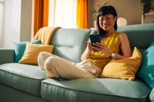 Happy asian woman using smartphone on green couch, vibrant living room